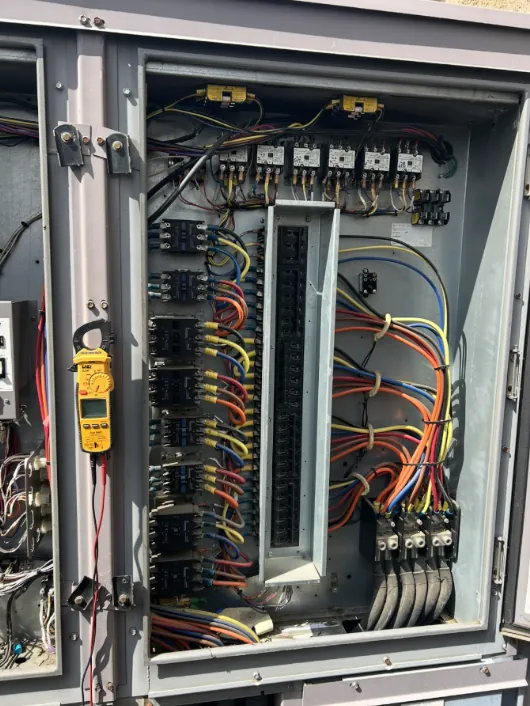 Clamp meter hanging on the door of an open control panel lined with contactors and wiring